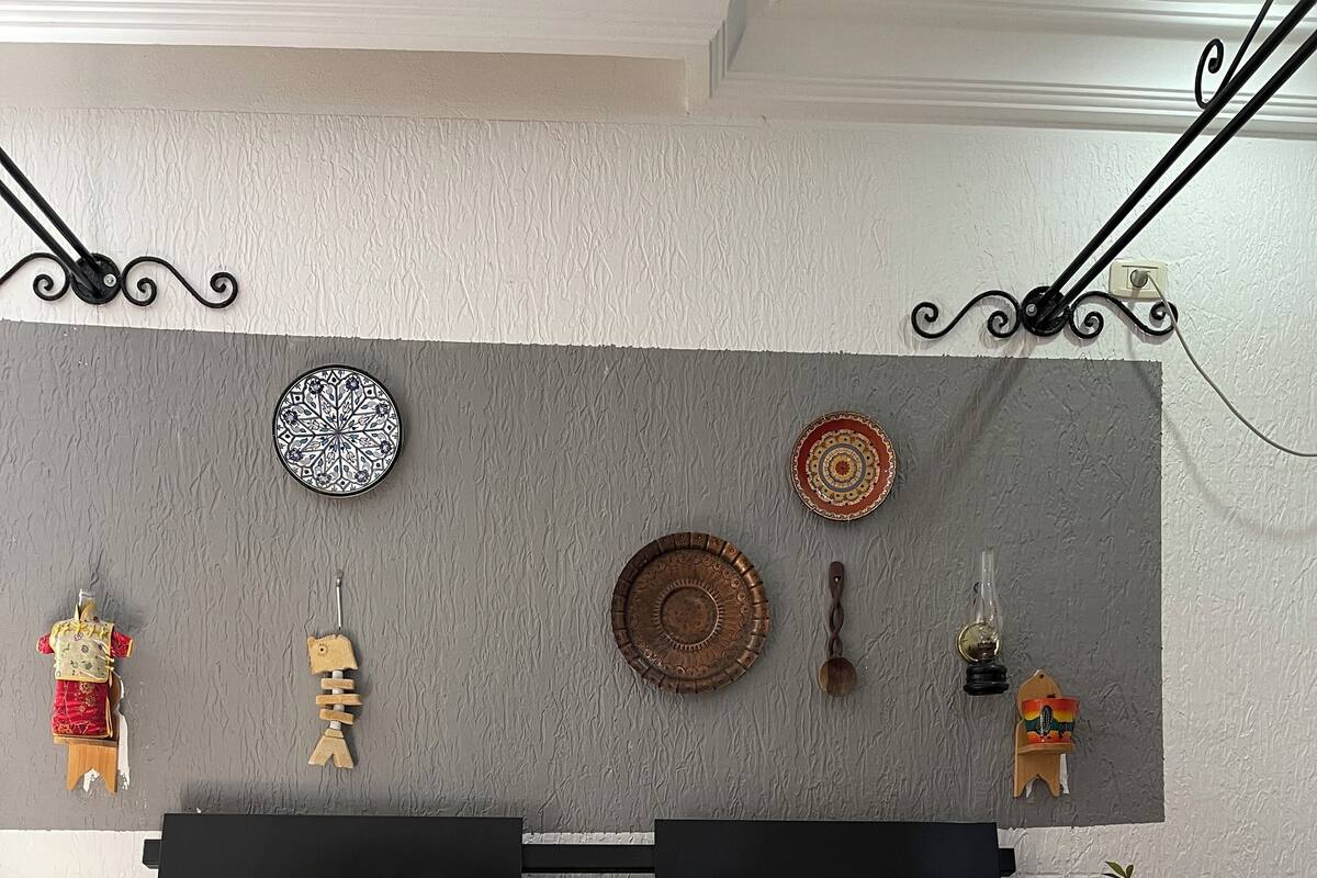 Una cama bien hecha con un edredón estampado, una almohada roja y amarilla, y una pared decorada con varios objetos, incluyendo un plato, un pez de madera y una pequeña planta.