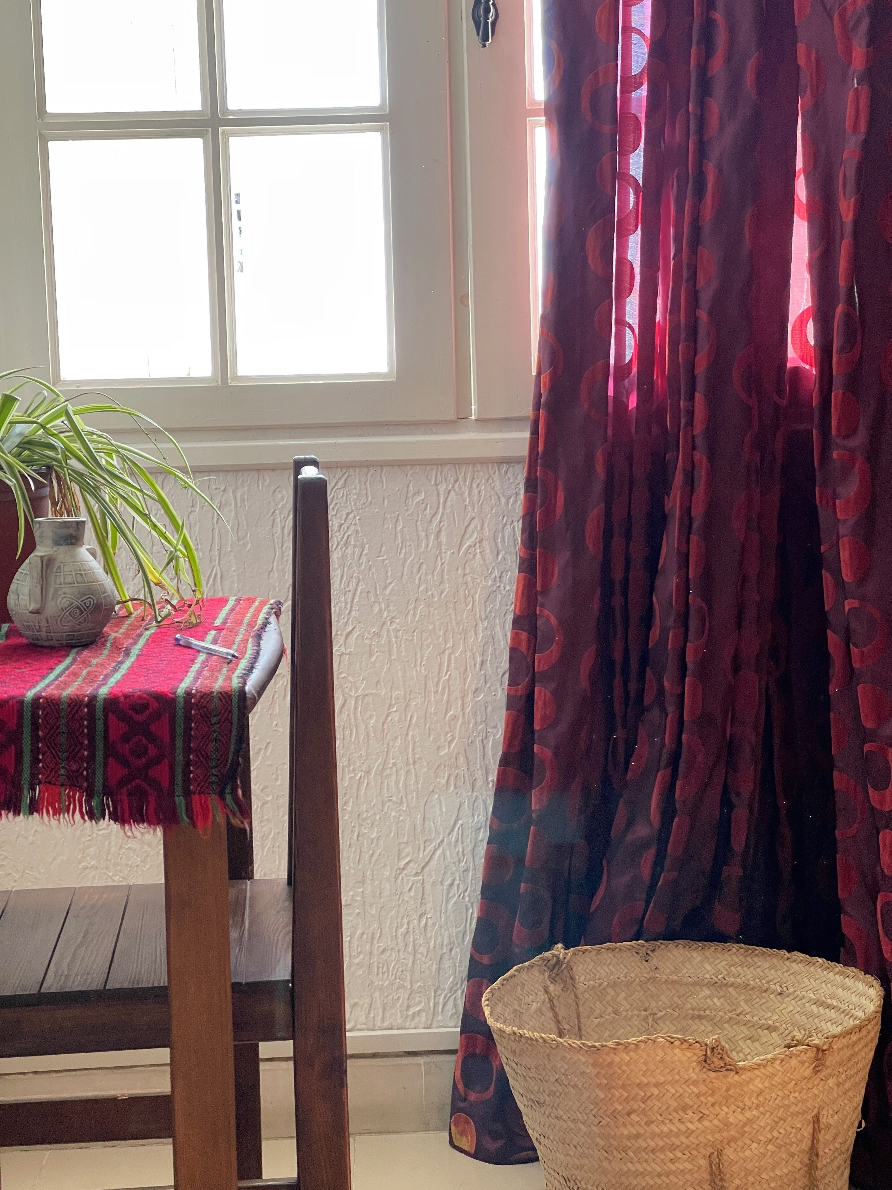 Una habitación con una mesa cubierta con un mantel estampado, una planta en maceta, una cesta tejida y una ventana con cortinas.