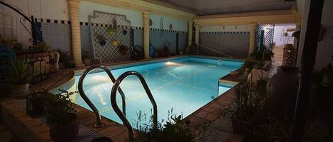 An indoor swimming pool with a tiled floor, surrounded by potted plants and a decorative railing.