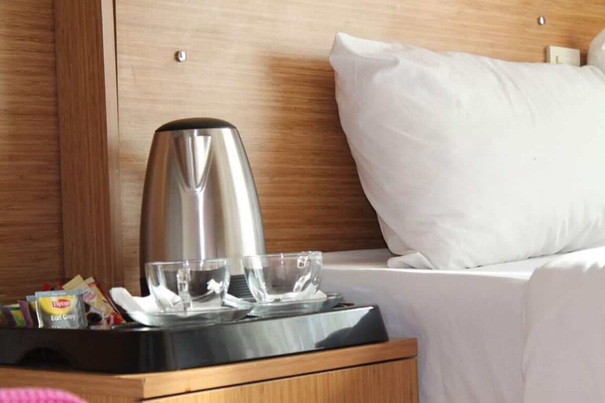 Una cama bien hecha con sábanas y almohadas blancas, una mesita de noche con una tetera de acero inoxidable, dos vasos y una bandeja con aperitivos.