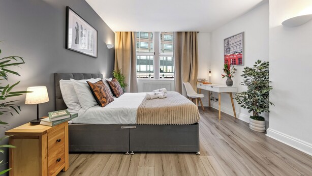 A modern bedroom with a bed, bedside table, lamp, and a view of the city through large windows. - Prime Liverpool Street Retreat (London)