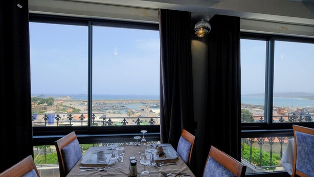A dining table set for four with a view of the sea and cityscape through large windows. - hotel baba aroudj  (Jijel)