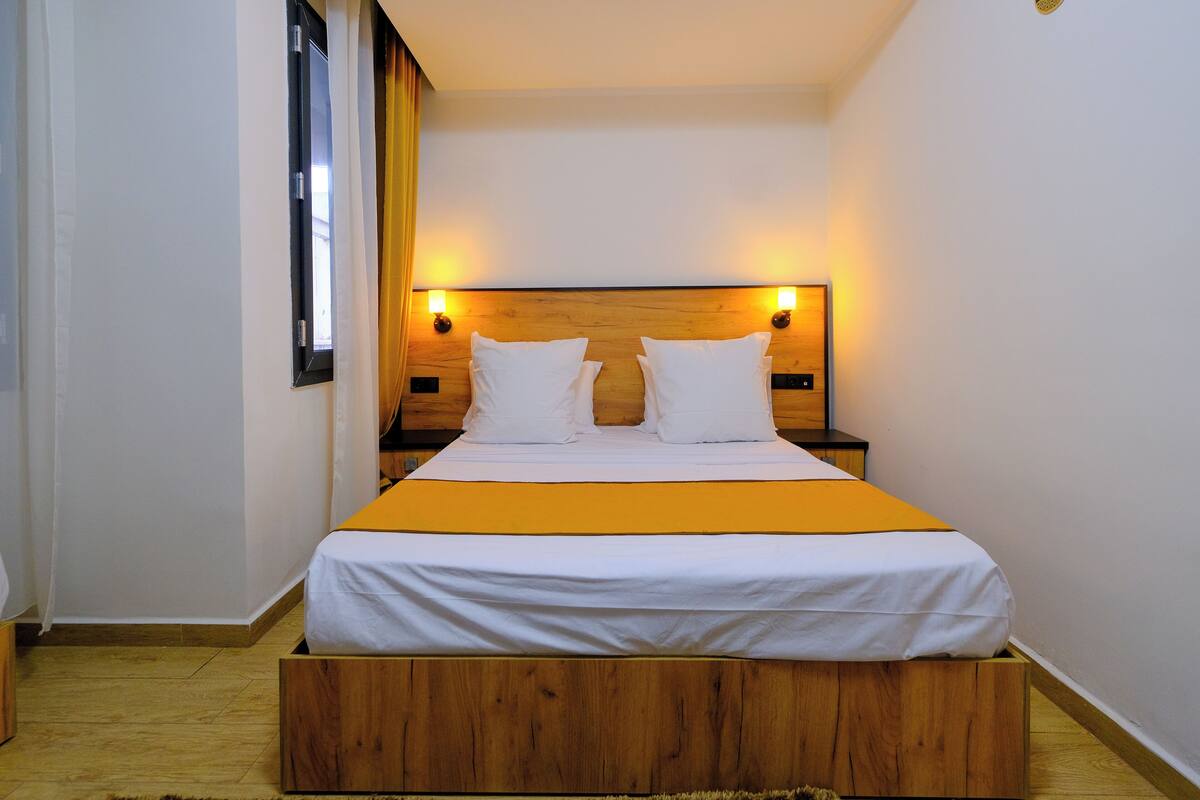 Ein Schlafzimmer mit einem hölzernen Kopfteil, zwei Nachttischen, einem Bett mit weißer Bettwäsche und einem gelben Akzent auf der Tagesdecke.