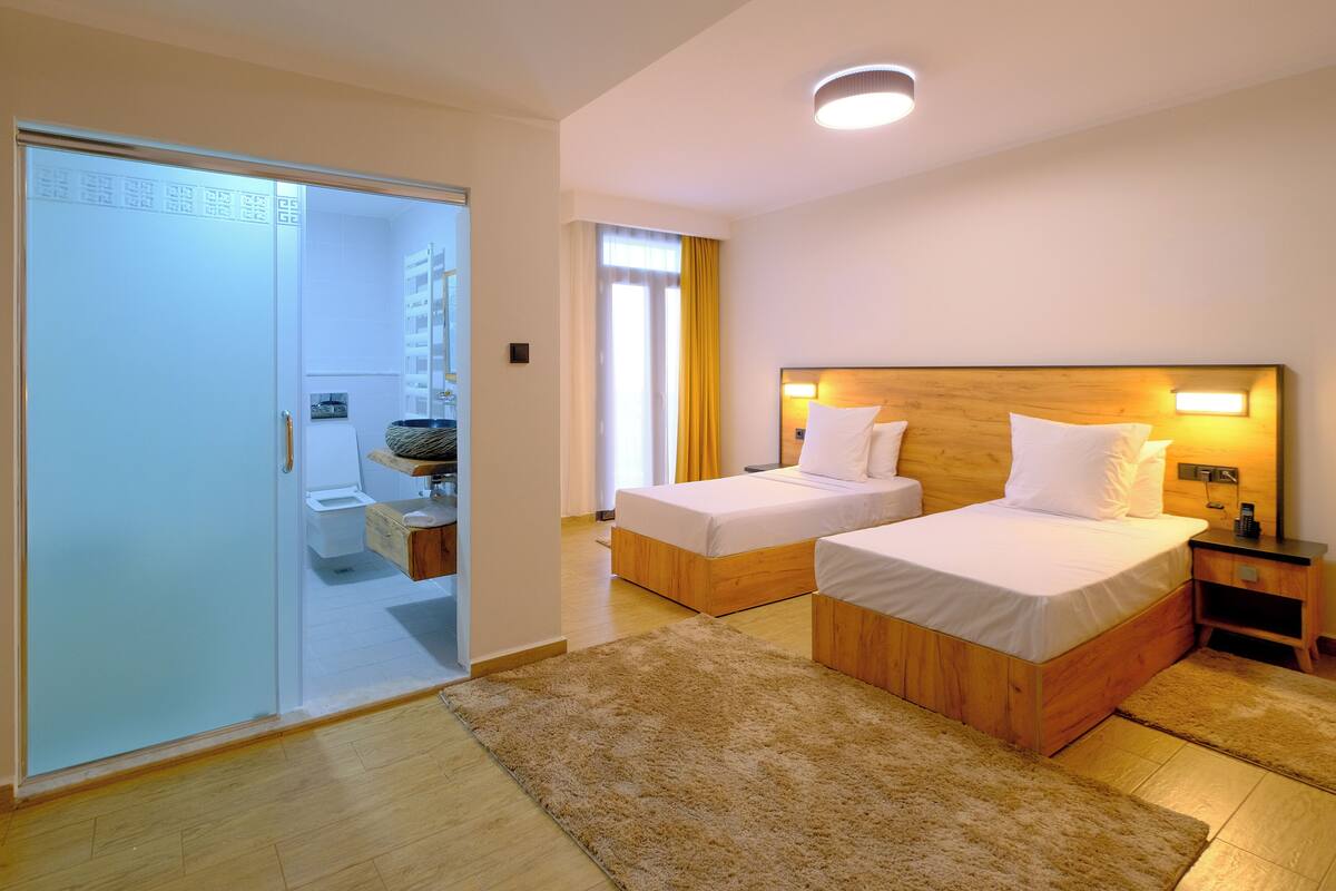 Ein Hotelzimmer mit zwei Einzelbetten, einem Holzkopfstück, einem Nachttisch, einem Bad mit Toilette und Waschbecken sowie einem Schiebetürenschrank.