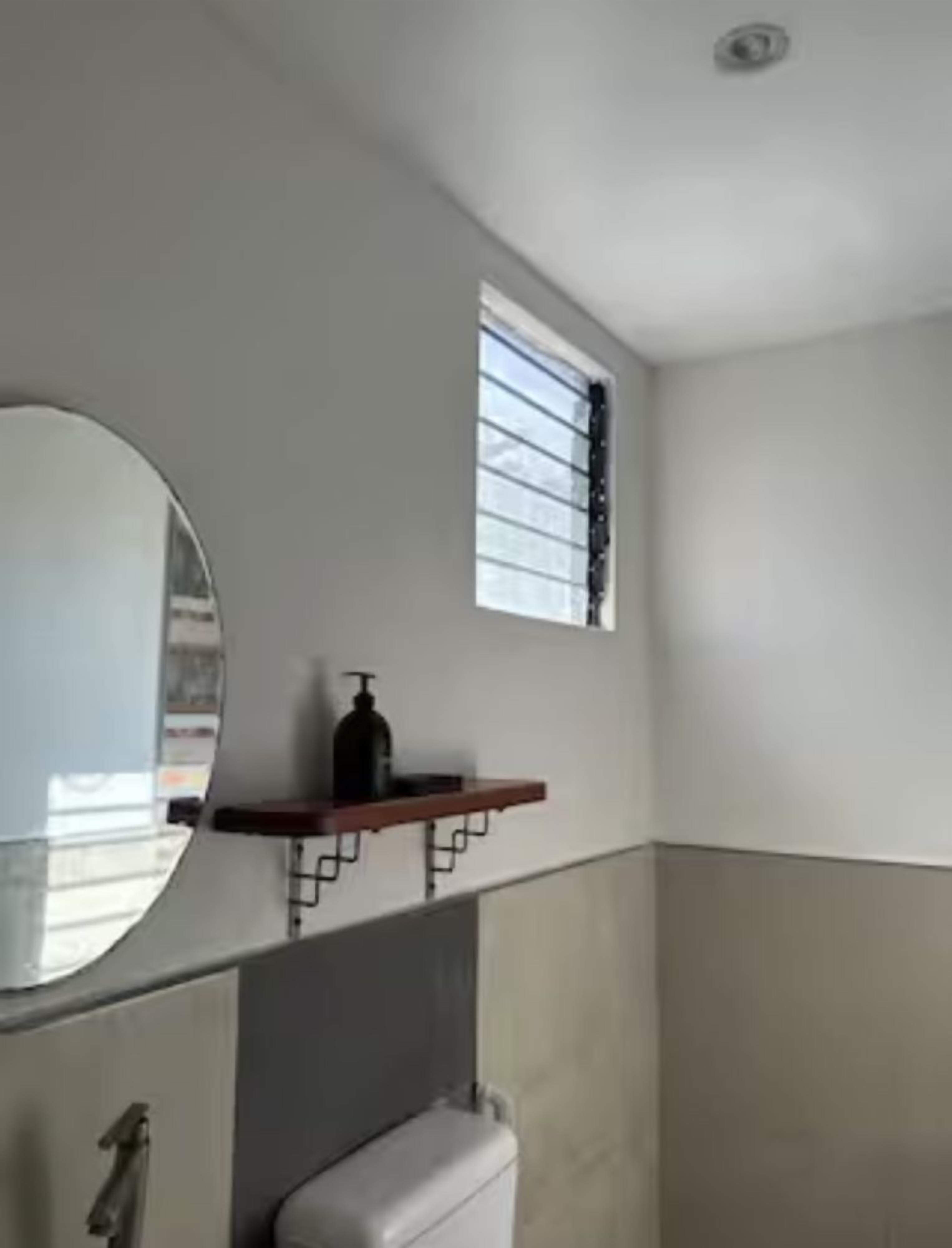 Un baño con inodoro, espejo, ventana y repisa con una botella.