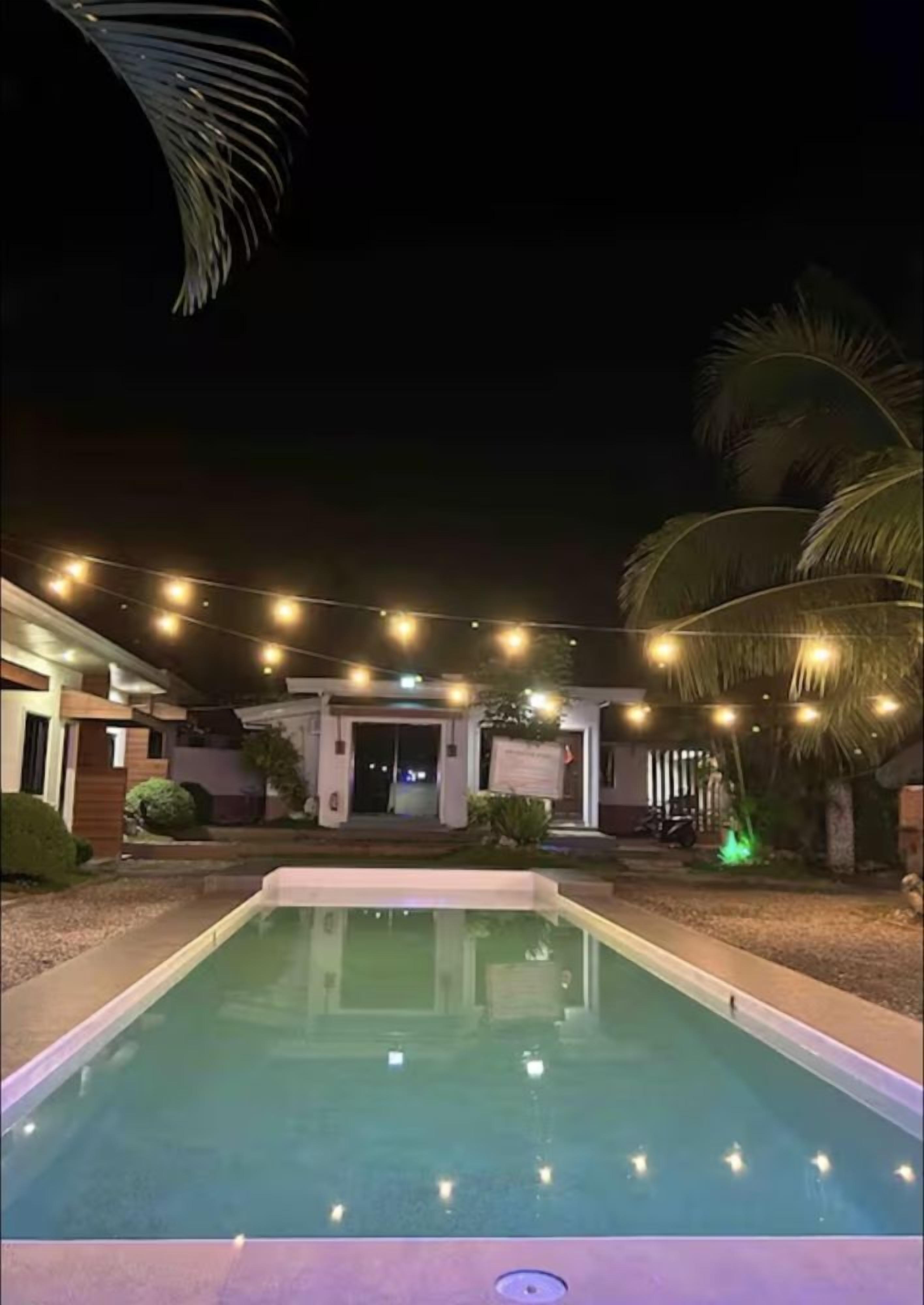 Une piscine décorée de guirlandes lumineuses, bordée de palmiers, et un bâtiment doté d’une entrée abritée.