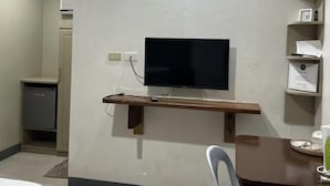 Ein Hotelzimmer mit Flachbildfernseher an der Wand, einem Holztisch mit Stuhl und einer kleinen Küchenzeile mit Kühlschrank und Spüle.