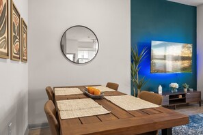 Un comedor con una mesa de madera, individuales y un bol de fruta. Hay un espejo redondo en la pared, un televisor de pantalla plana y una planta sobre un atril.