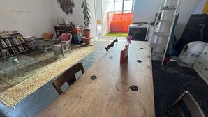 Een eethoek met een houten tafel, stoelen en een rode vaas. Een boekenplank met boeken en een bloempot zijn te zien. Achtergrond: een ladder en een witte kast.