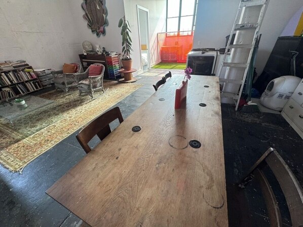 A dining area with a wooden table, chairs, and a red vase. There is a bookshelf with books and a potted plant. A ladder and a white cabinet are visible in the background.