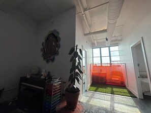 A room with a large window, a potted plant, a desk with drawers, and a red safety net in the background.