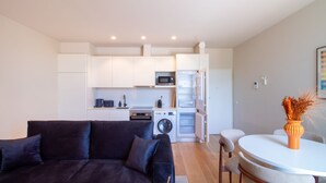 Un espacio habitable compacto con un sofá de color oscuro, una pequeña mesa de comedor y una kitchenette con microondas, horno y lavadora.
