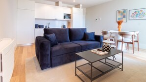 Una sala de estar moderna con un sofá azul oscuro, una mesa de centro y una zona de cocina con armarios y electrodomésticos blancos.