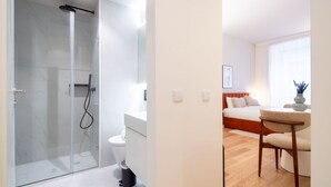 Moderni hotellihuone, jossa on lasiseinällä varustettu suihku, valkoinen wc-istuin, puulattia, valkoisilla lakanoilla varustettu sänky, yöpöytä ja tuoli.