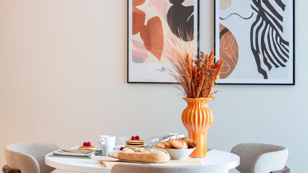 Et spisebord med morgenmadsanretninger, en vase med tørrede planter og abstrakt kunst på væggen.