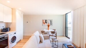En moderne lejlighed med et køkken, spiseområde og opholdsstue. Der er vaskemaskine, spisebord med stole, sofa og et stort vindue med gardiner.