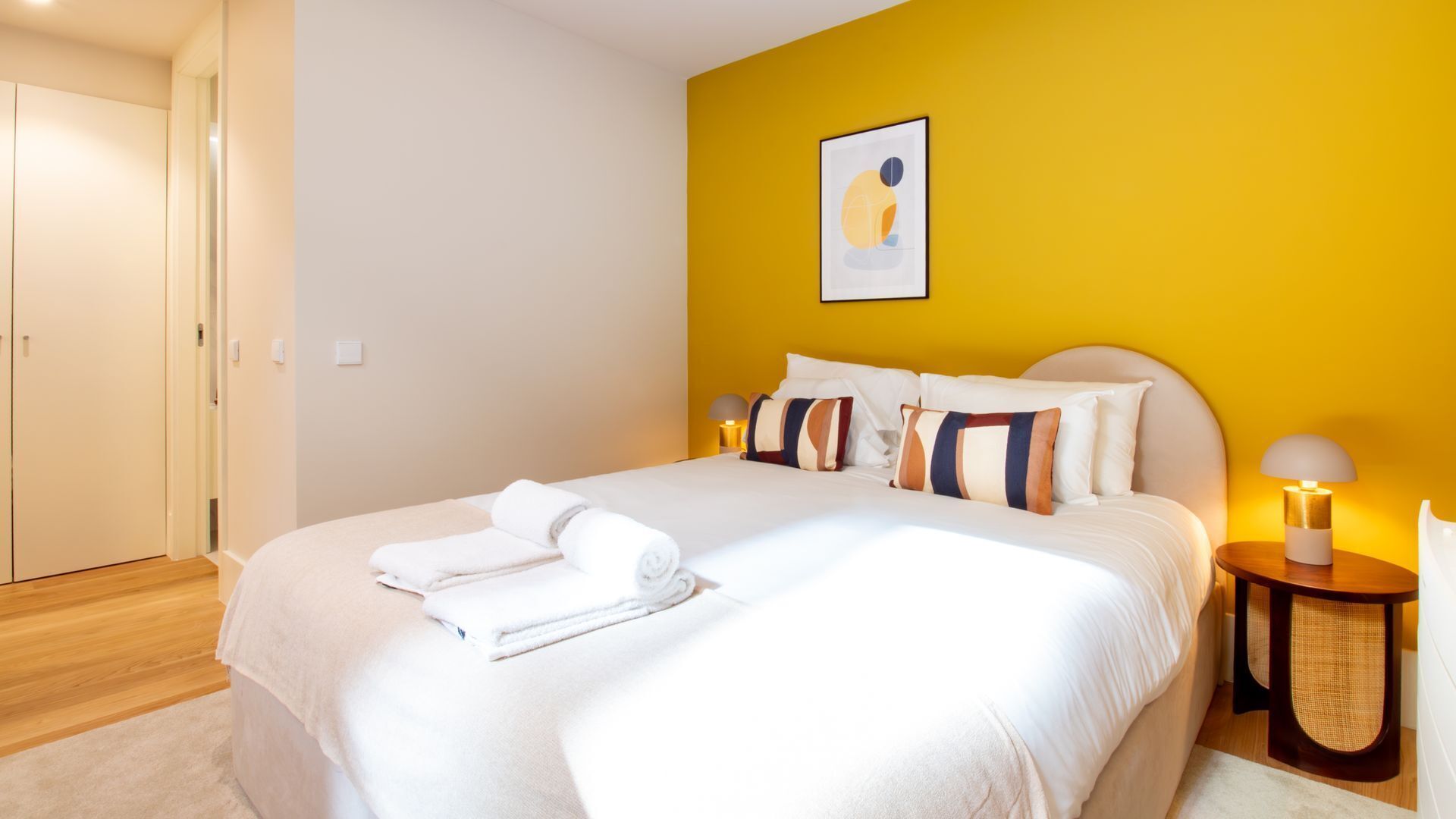 A neatly made bed with white linens and rolled towels, a bedside table with a lamp, and a wall with a framed abstract artwork.