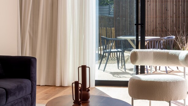 Ein modernes Wohnzimmer mit einem dunklen Sofa, einem runden Tisch und einem weißen Pouf. Ein großes Fenster mit weißen Vorhängen führt auf eine Außenpatio mit Tisch und Stühlen.