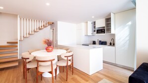 Eine moderne Küche mit weißen Schränken, einer Mikrowelle und einer Kaffeemaschine. Ein Essbereich mit weißem Tisch und Stühlen sowie eine Treppe mit Holzstufen.