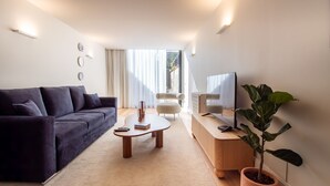 Ein modernes Wohnzimmer mit einem dunklen Sofa, einem Holztisch, einem Fernseher auf einem Holzständer und einer großen Kühlpflanze.