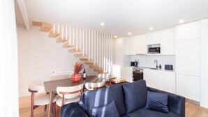 A modern kitchen with white cabinets, a microwave, and a stove. There is a dining area with a table and chairs, and a blue sofa in the living room. - Miguel Bombarda House - 402 by LovelyStay (Porto)