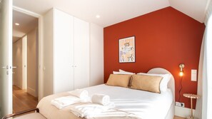Ein Schlafzimmer mit Bett, weißen Wänden, einer roten Akzentwand, einem Gemälde, einer Nachttischlampe und einer Stehlampe.