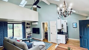 Een ruime woonkamer met een hoge nok, een plafondventilator, een kroonluchter, een grijze bank, een eettafel en een keukenhoek.