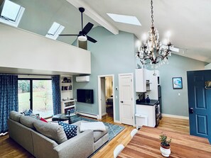 A spacious living area with a high ceiling, a ceiling fan, a chandelier, a grey sofa, a dining table, and a kitchen area.
