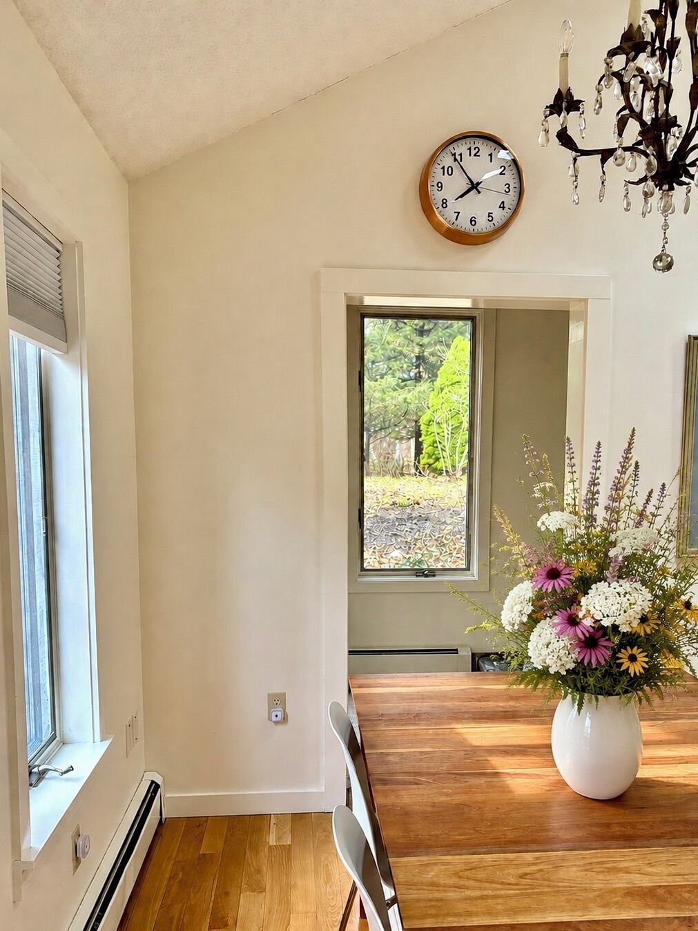 A dining area with a wooden table, chairs, a clock, and a window with a view of greenery.