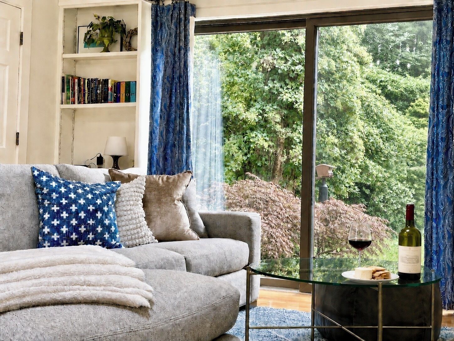 A living room with a grey sofa, patterned cushions, a glass coffee table with a bottle and glasses, and a bookshelf with books and decorative items.