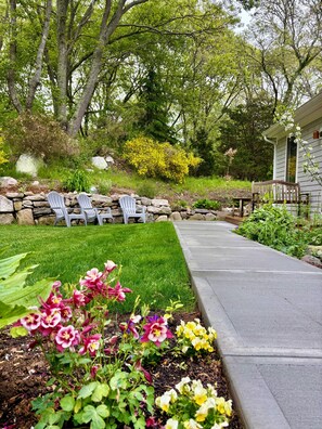 A garden with a stone wall, Adirondack chairs, and a pathway leading to a house.