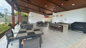 Un espace extérieur aménagé pour les repas, avec des tables et des chaises, un barbecue et des ventilateurs de plafond.