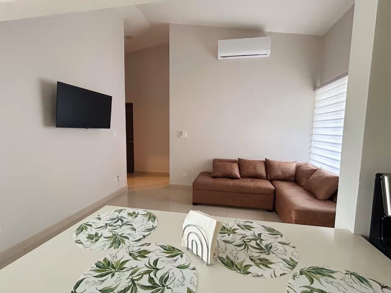 A modern living room with a sofa, a flat-screen TV mounted on the wall, and a table with a floral tablecloth.