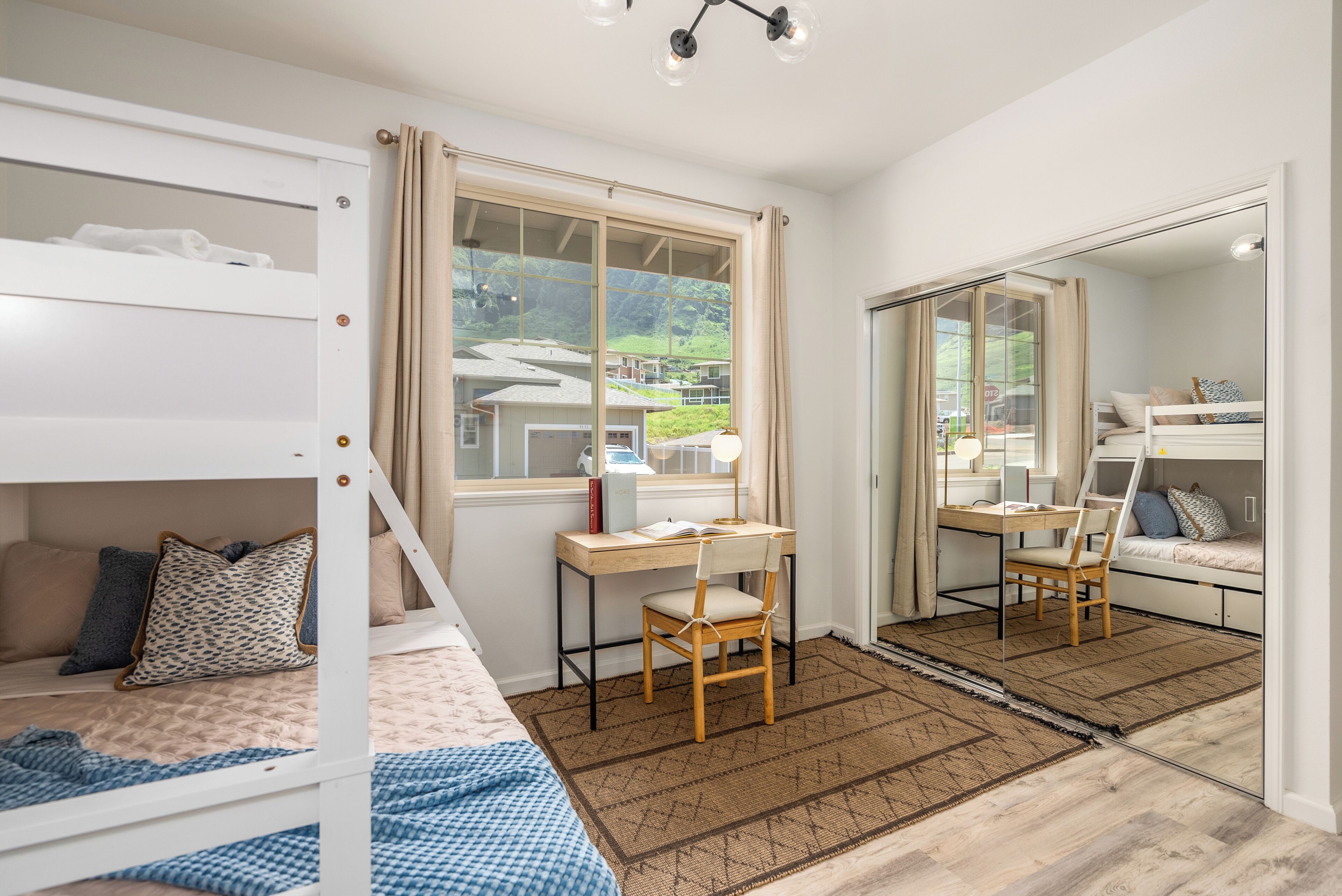 A bedroom with a bunk bed, a desk with a chair, a large window, and a mirror.