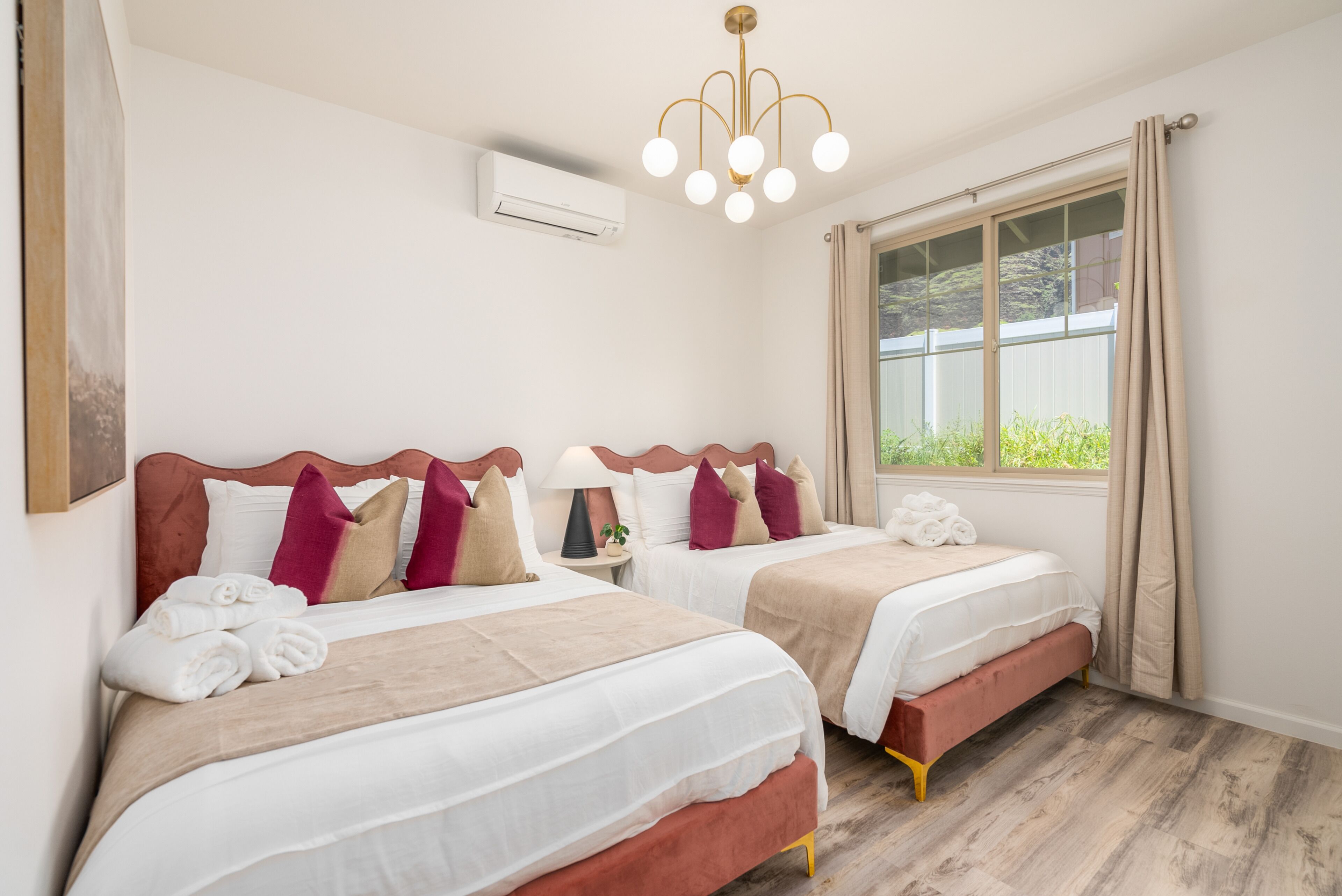 A hotel room with two beds, a chandelier, an air conditioner, and a window with curtains.