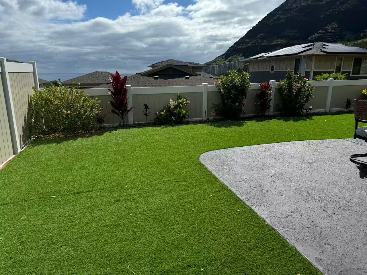 Une pelouse bien entretenue avec une terrasse en béton, entourée d’une clôture en bois et agrémentée de diverses plantes.