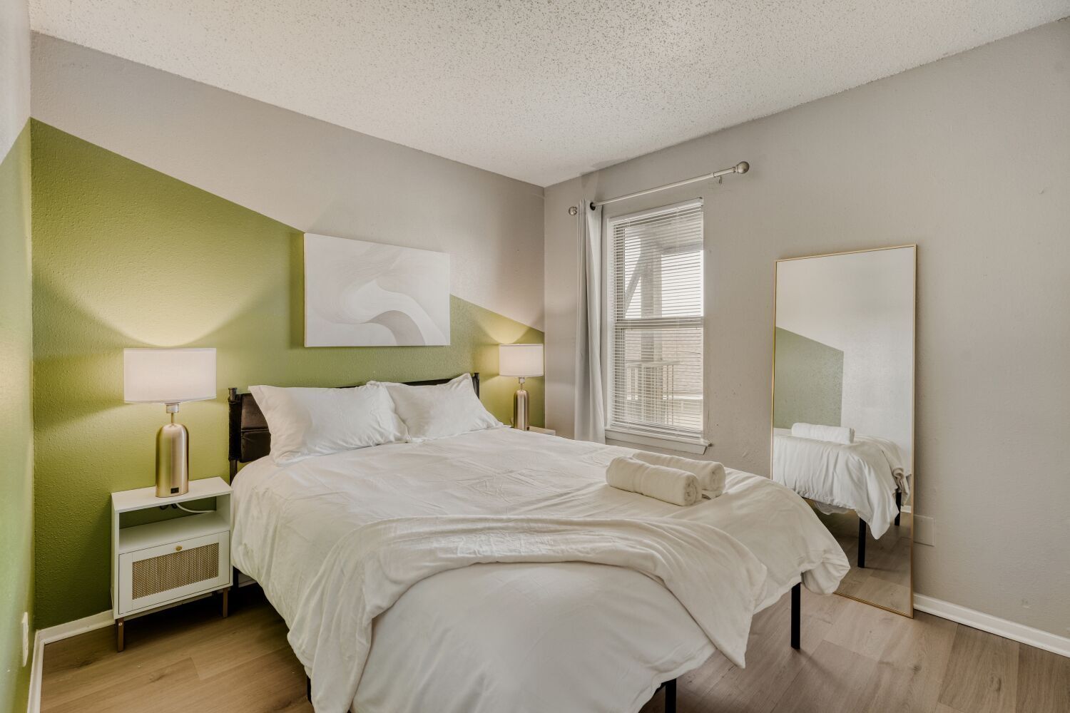 Una cama bien hecha en una habitación con un muro verde decorativo, una lámpara de noche y un espejo grande.
