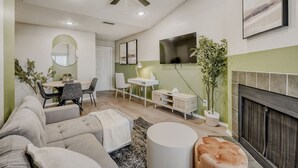 A living room with a sofa, a dining table with chairs, a TV mounted on the wall, a fireplace, and a ceiling fan. - Urban Chic Getaway 2B | Sleeps 6 | Pool, Prkg, W/D (Austin)