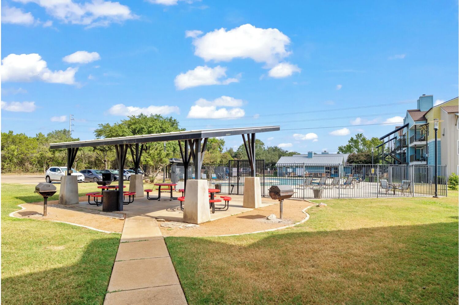 Un área de picnic techada con mesas y parrillas, rodeada por una piscina cercada y un edificio con balcón.