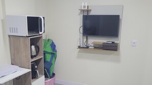 A small, neatly organized room with a wooden table, a white chair, a microwave, a television, and a shelf with various items. - Furnished one-bedroom apartment in Balneário Gaivota, right in the center. (Balneário Gaivota)
