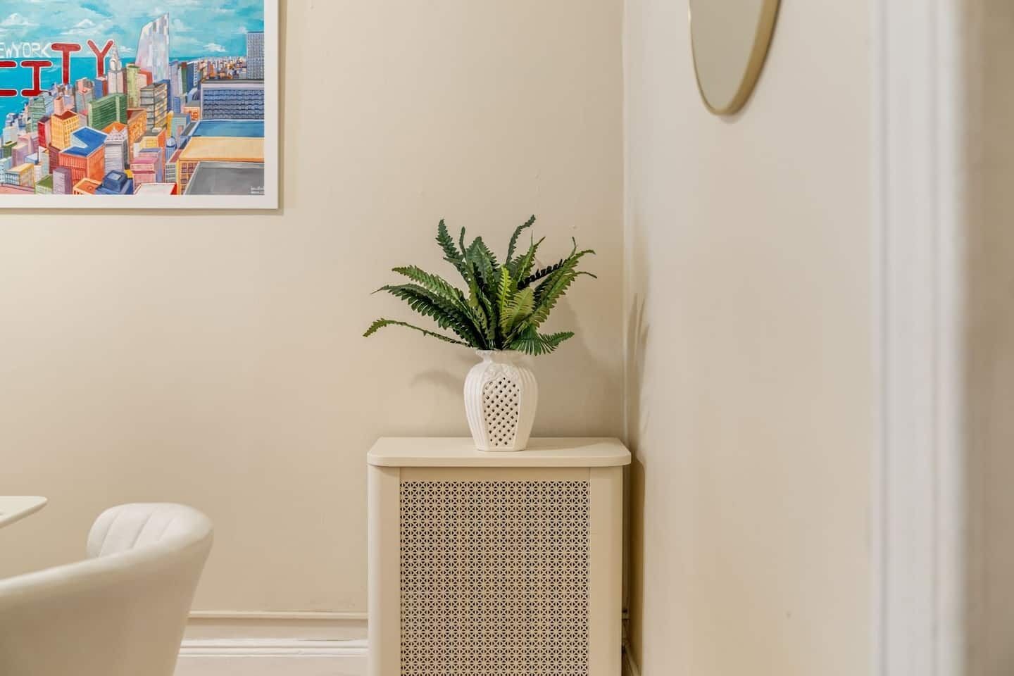 A room with a white radiator, a potted plant, and a framed cityscape painting on the wall.