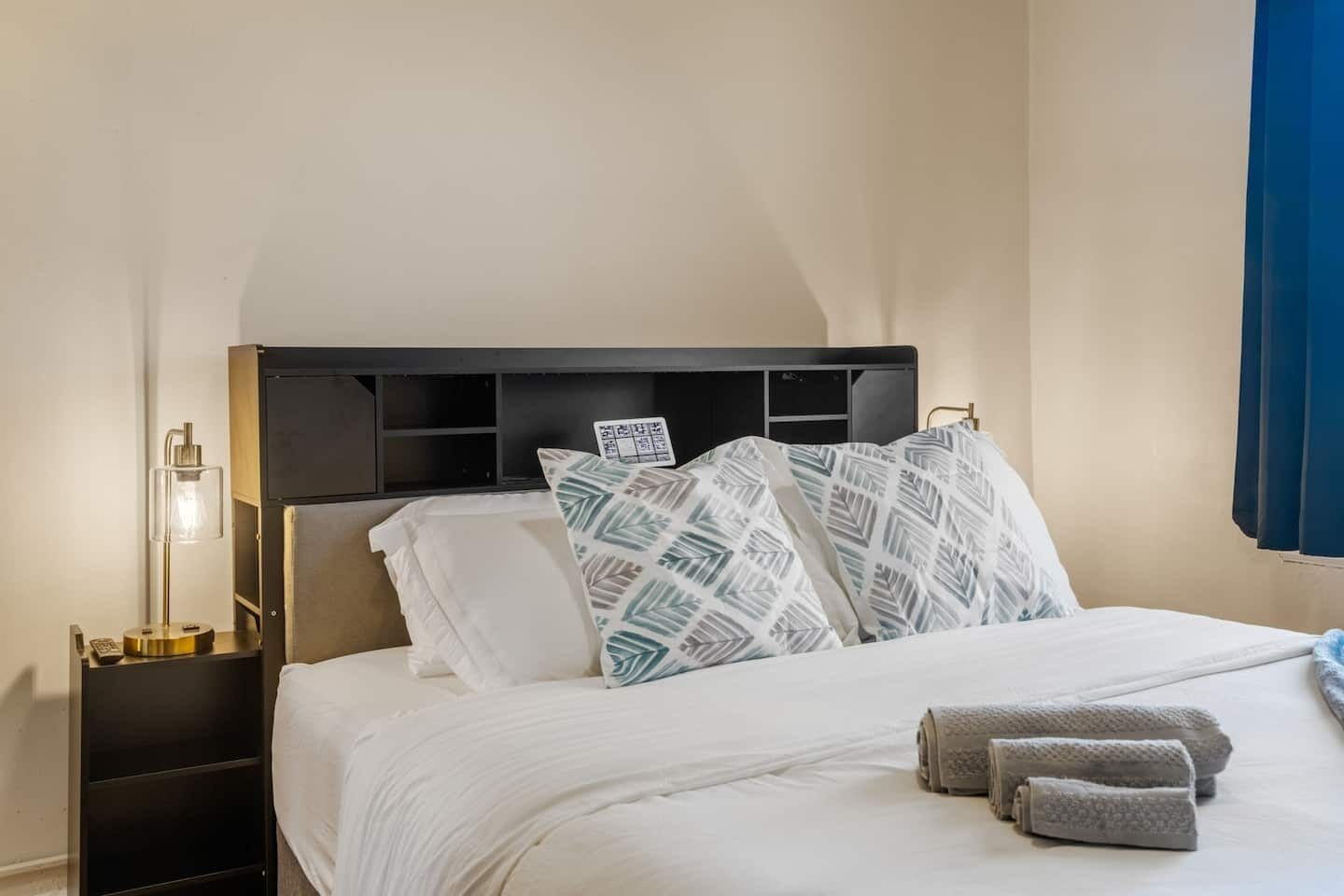 A neatly made bed with patterned pillows, a bedside lamp, and rolled towels on a nightstand.