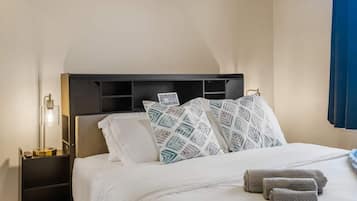 A neatly made bed with patterned pillows, a bedside lamp, and rolled towels on a nightstand.