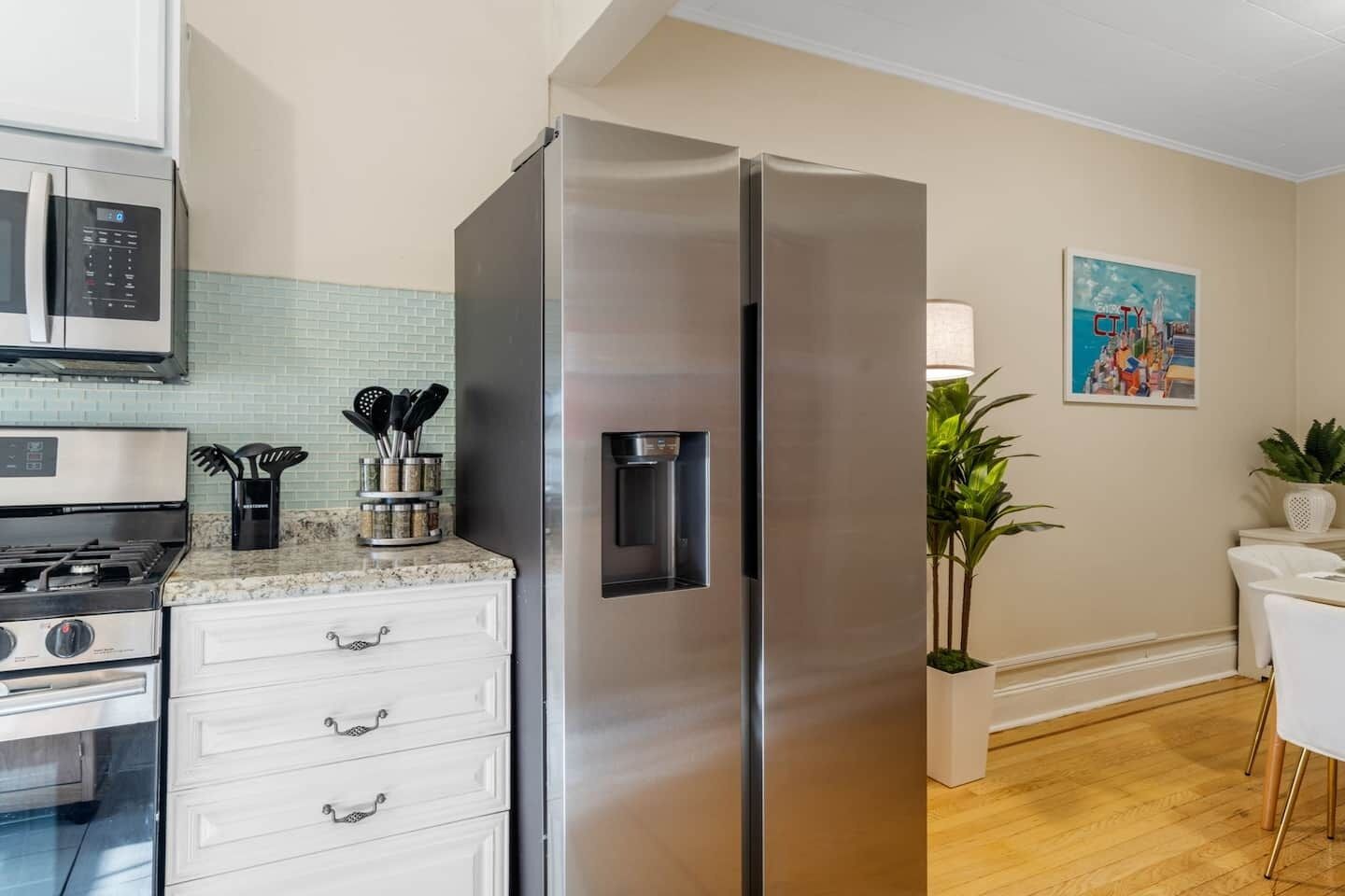 A kitchen with a stainless steel refrigerator, a microwave, a stove, and a sink. There is a plant and a painting on the wall.