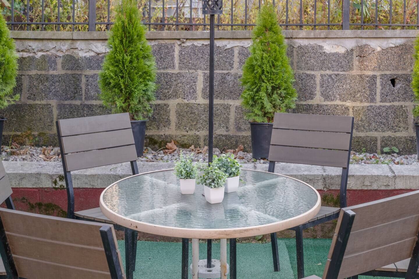 A round glass table with four chairs, three potted plants, and a stone wall with a metal fence in the background.
