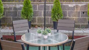 A round glass table with four chairs, three potted plants, and a stone wall with a metal fence in the background.