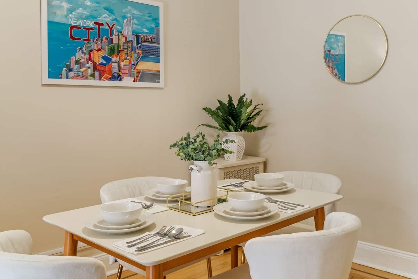 A dining area with a white table set for four, a cityscape painting, a potted plant, and a round mirror on the wall.