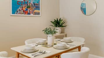 A dining area with a white table set for four, a cityscape painting, a potted plant, and a round mirror on the wall.