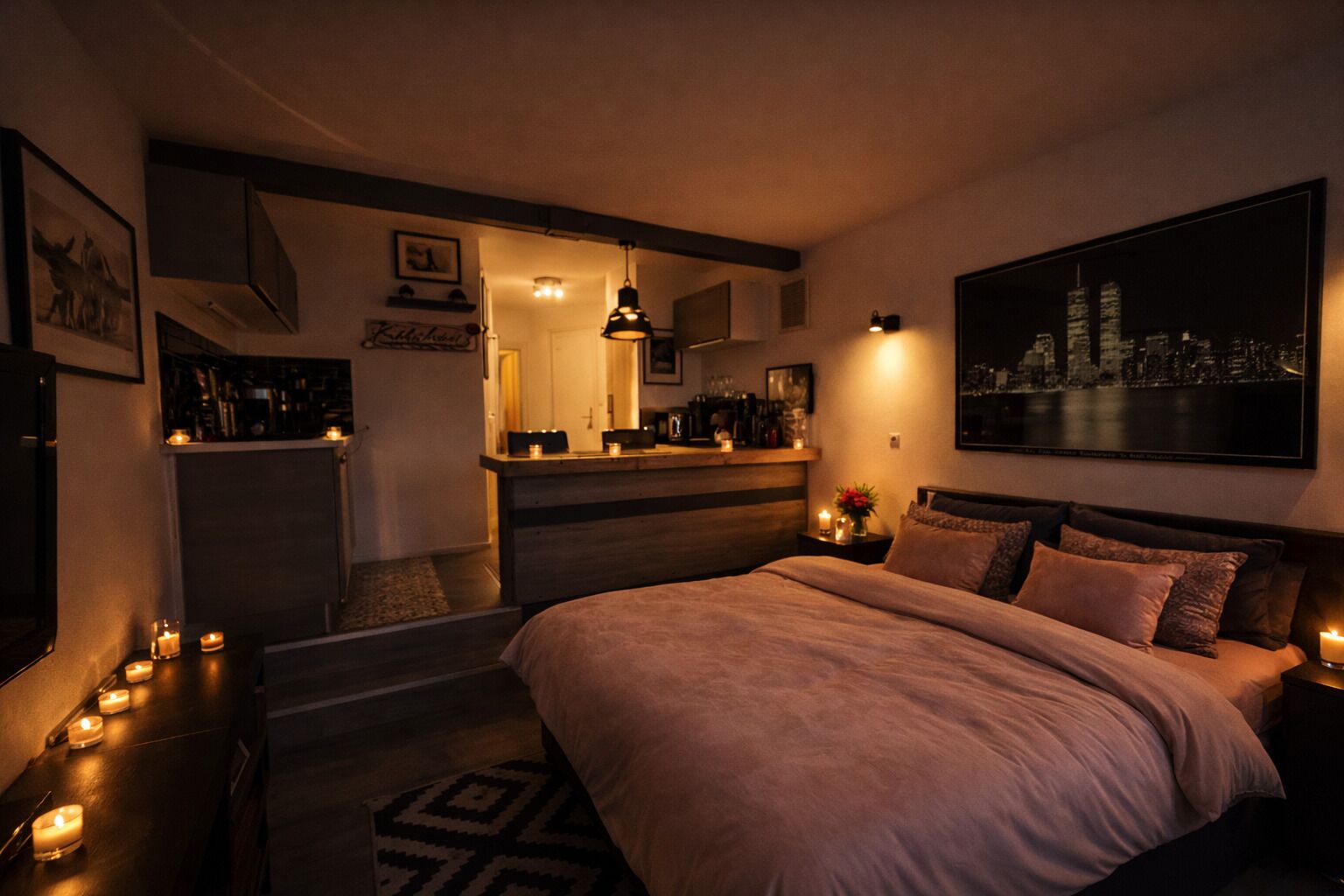 Une chambre confortable avec un grand lit, des tables de chevet ornées de bougies, une table de nuit, un tableau représentant un paysage urbain et, au fond, une petite cuisine.