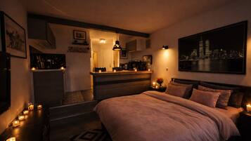 Une chambre confortable avec un grand lit, des tables de chevet ornées de bougies, une table de nuit, un tableau représentant un paysage urbain et, au fond, une petite cuisine.